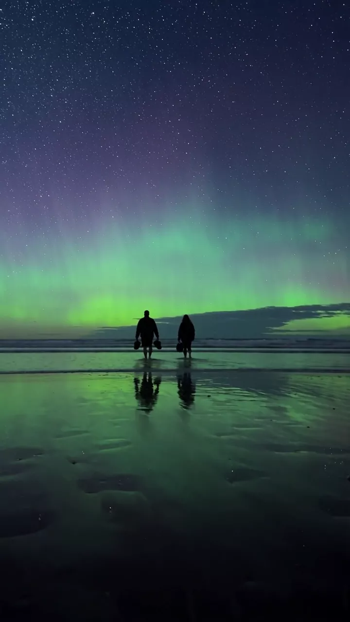 A couple wades along a wet beach at dusk under a plain blue sky; a green and violet aurora fades in and fills the sky and its reflection in the sand