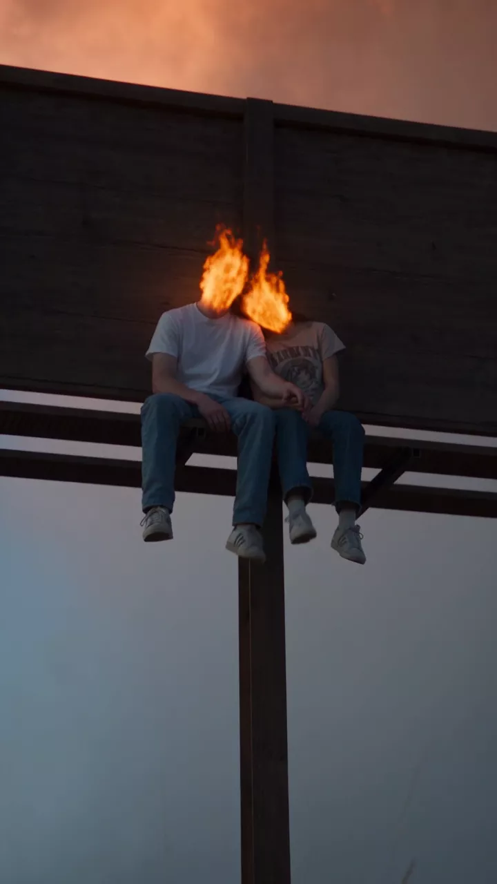 A couple with flames for heads sits on the catwalk of a roadside billboard under a towering orange-lit smoke cloud at blue hour