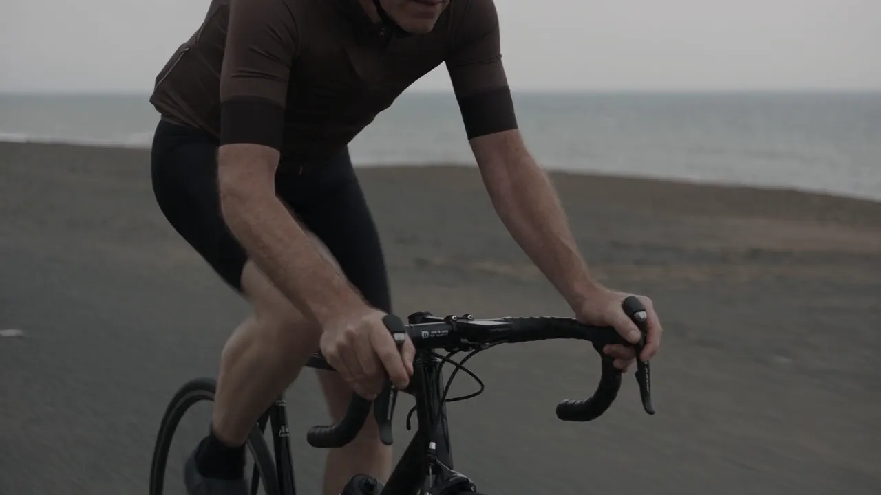 A cyclist in a brown jersey rides a road bike along a flat grey coastal road under an overcast sky, the sea behind him, in flat ungraded colour.
