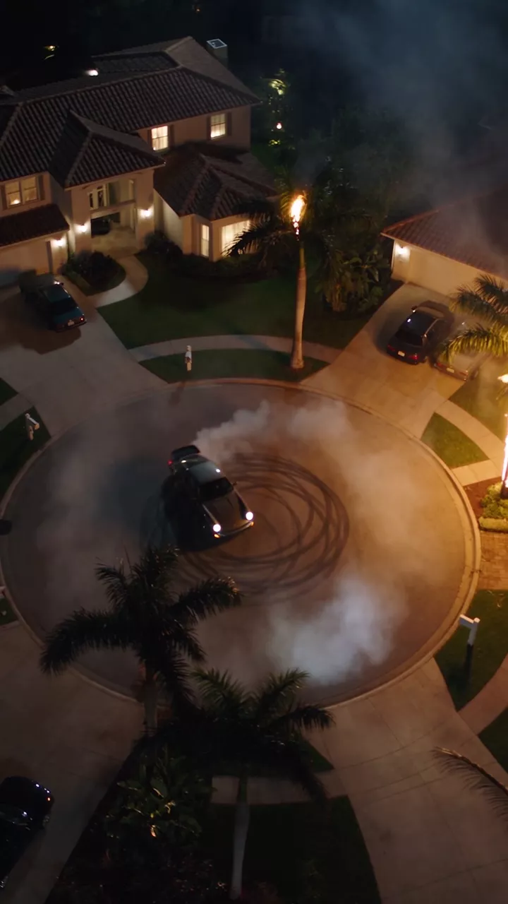 A black Porsche 911 spins donuts at a palm-lined Miami intersection at night, filmed low from the street through tire smoke with palm trees burning behind, then from high overhead as the car draws a circle of smoke