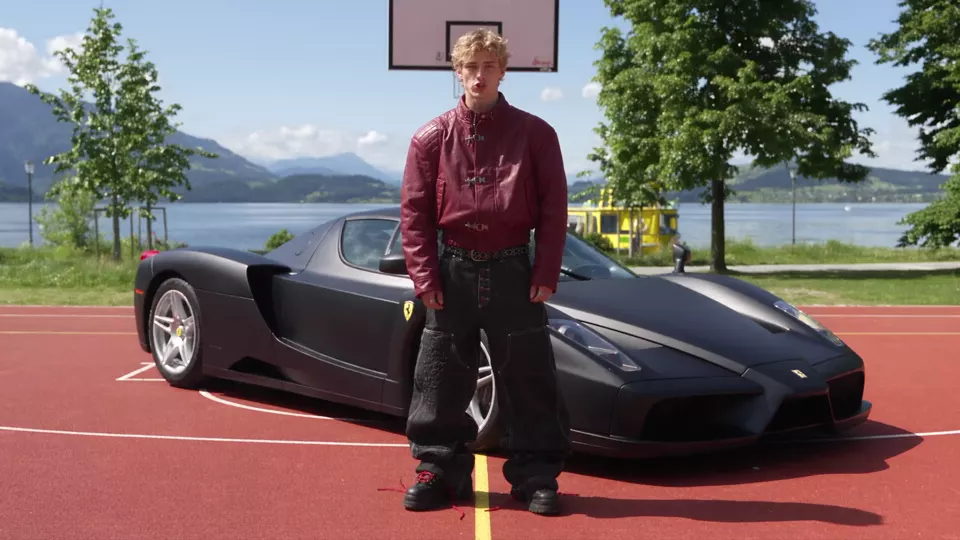 The take: a young man in a burgundy leather jacket stands beside a black Ferrari on a red basketball court by a lake, talks to the camera and turns his head up, left and right while the camera stays still