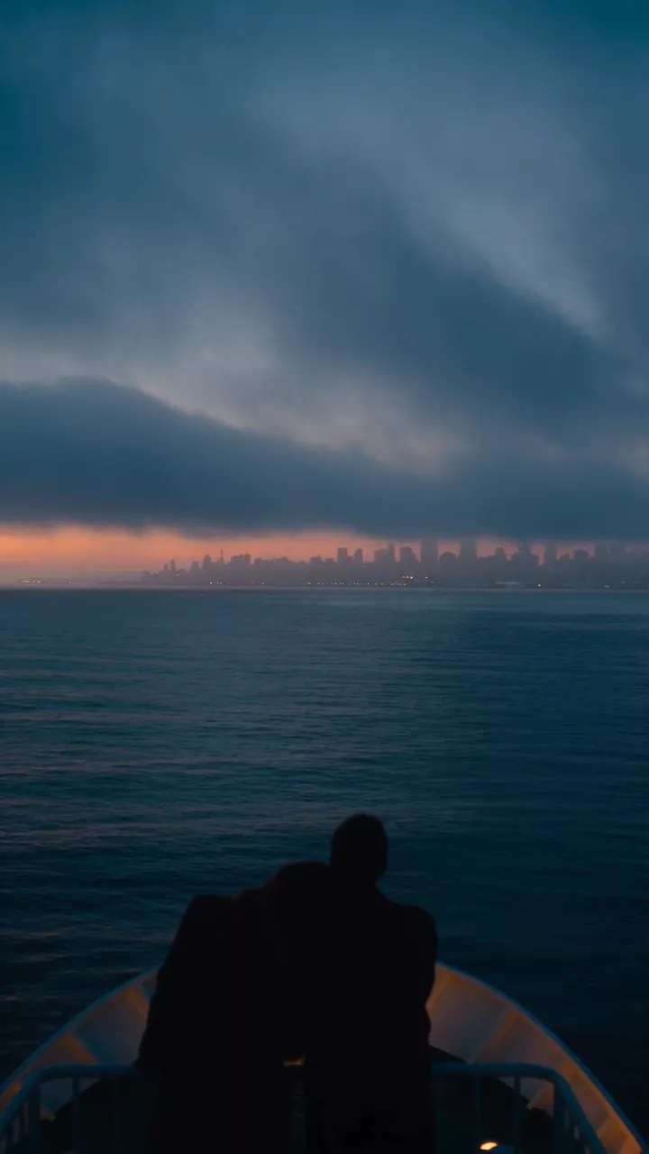 A couple sits at the back of a ferry at dusk while a city skyline fades into fog over the water