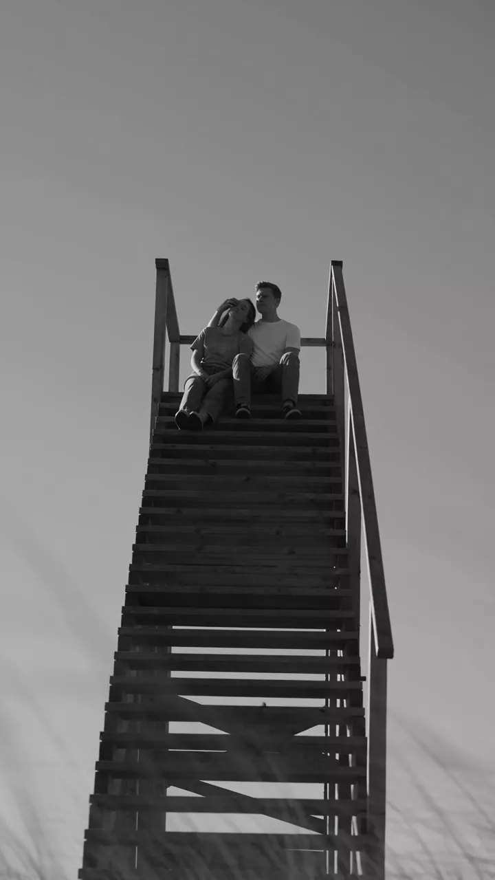 Black and white: a couple sits together at the top of a tall freestanding staircase in a grassy field, seen from below