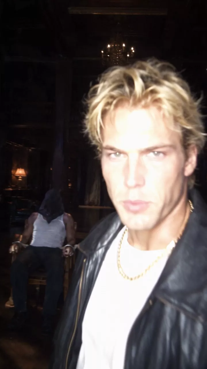 A dark mansion lit by a camera flash: a blond man in a black leather jacket and gold chain turns toward the camera and talks, then the camera pans to a blonde woman on a black leather couch petting a black panther