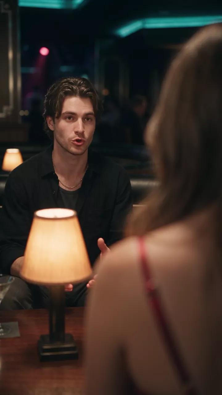 Over the woman's bare shoulder, with a table lamp in the foreground, the man across the booth explains something with a small hand gesture