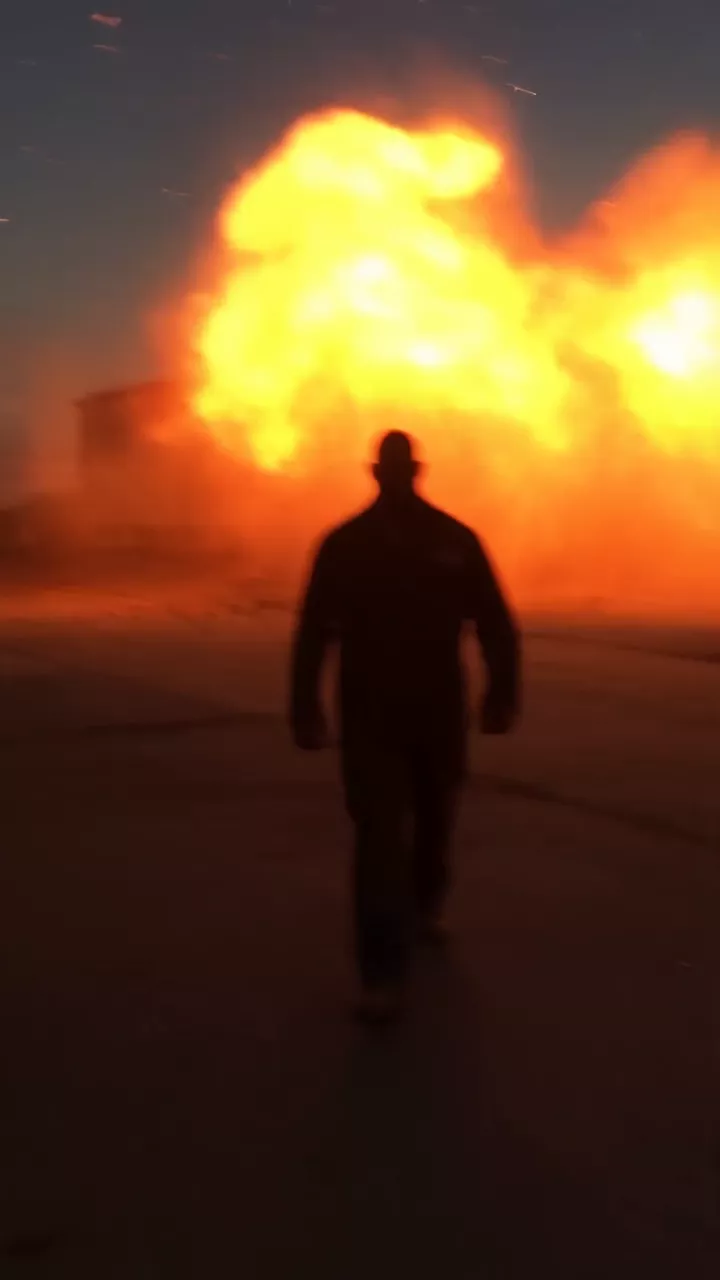 The same parking lot prompt run a second time: a man in work clothes walks toward the camera through a lot at dusk as a fireball goes up behind him, and he keeps coming until he fills the frame